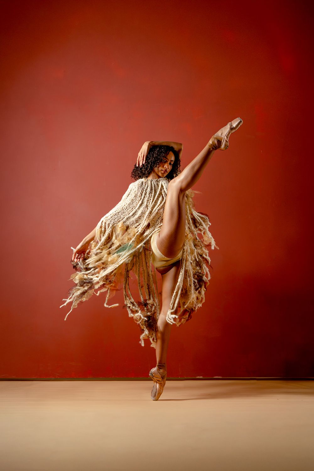 A ballet dancer in a fringed costume performs an arabesque on pointe against a red background.