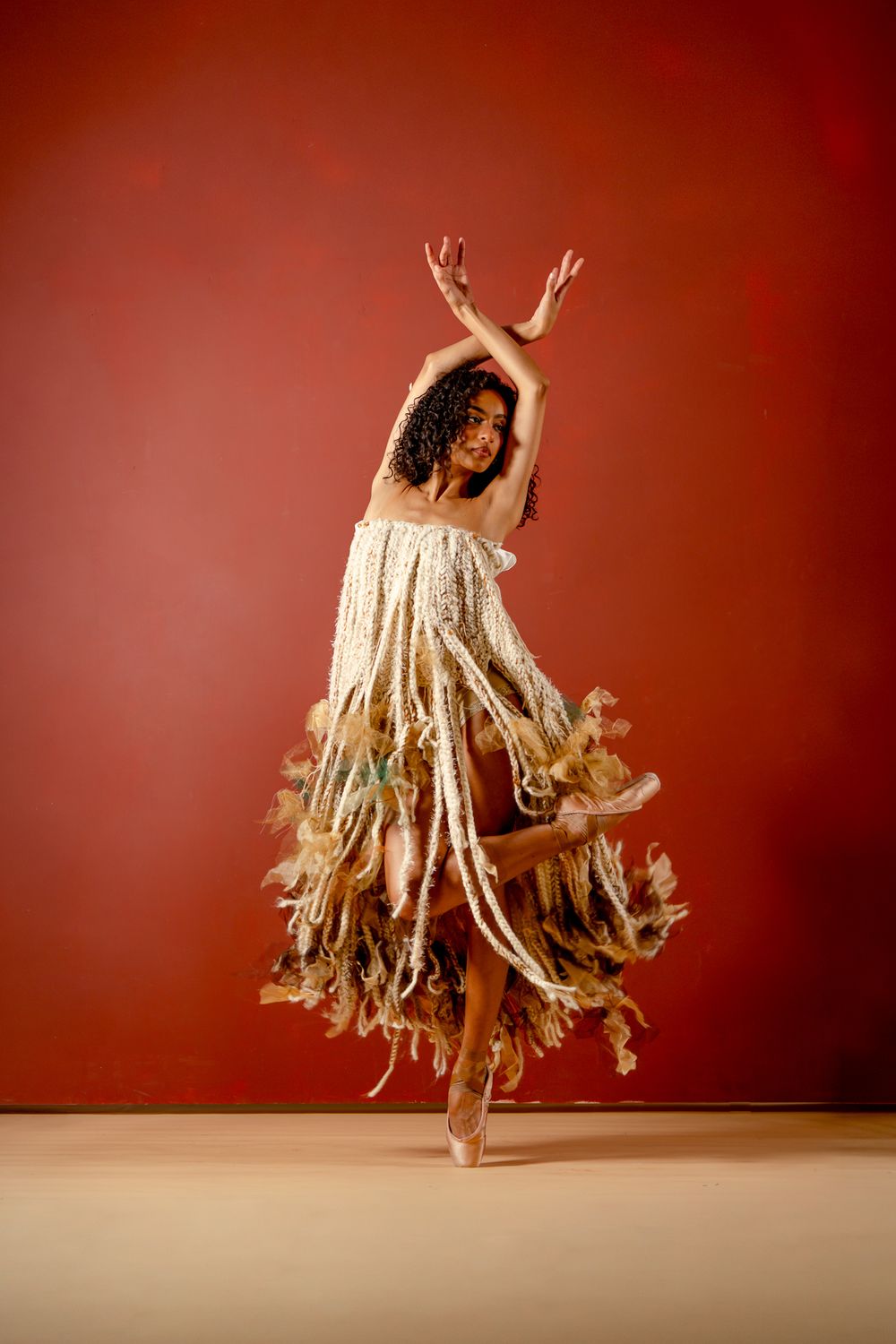 A dancer in a flowing fringed dress leaps gracefully with arms raised against a deep red background.