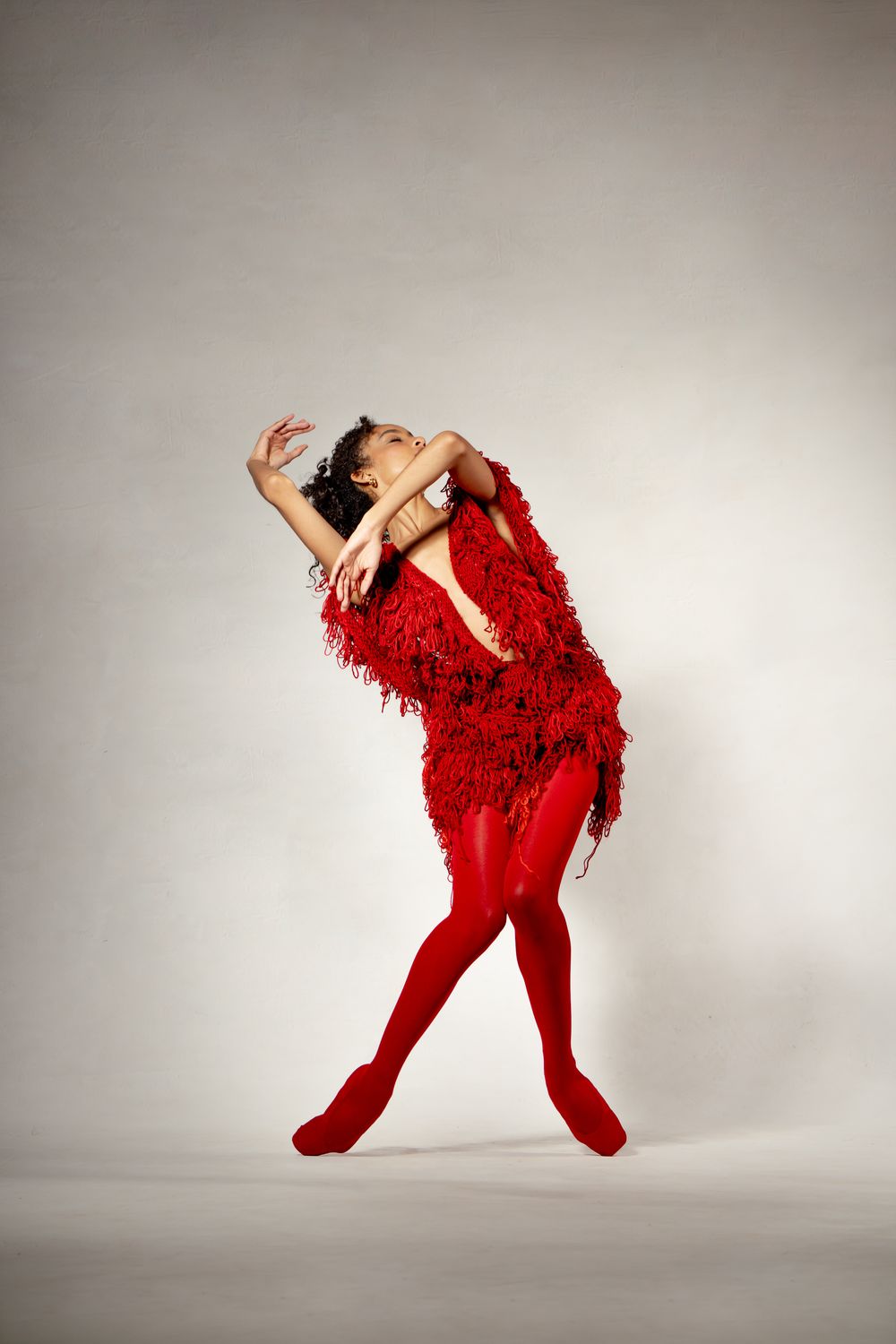 A dancer in a red fringed dress and red tights poses gracefully against a white background.