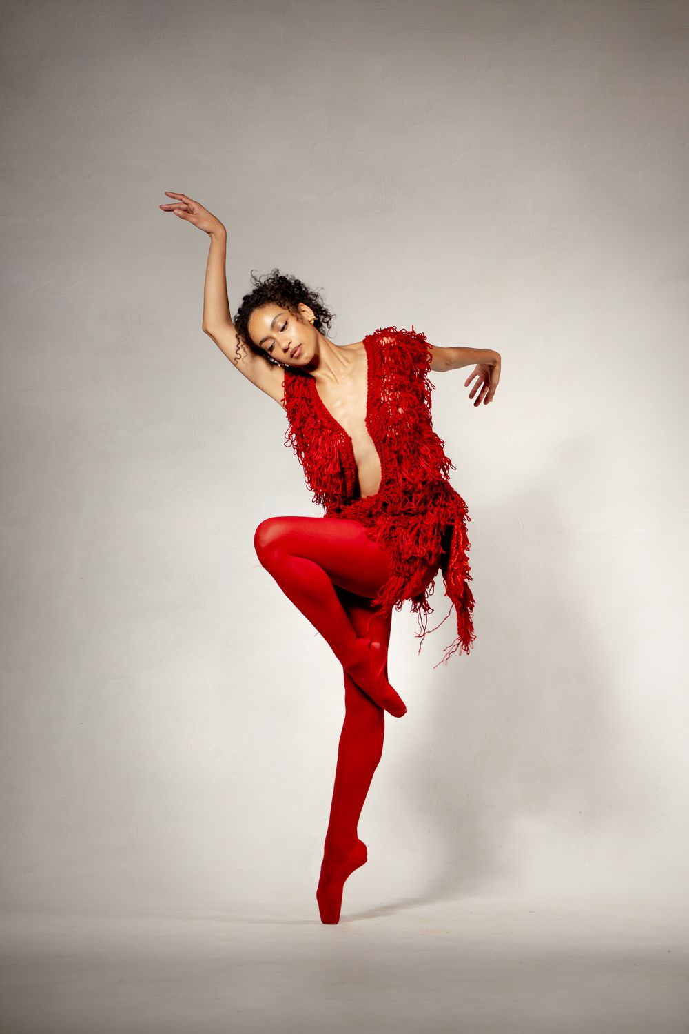A ballet dancer in a vibrant red fringed costume and tights performs a graceful leap on pointe.