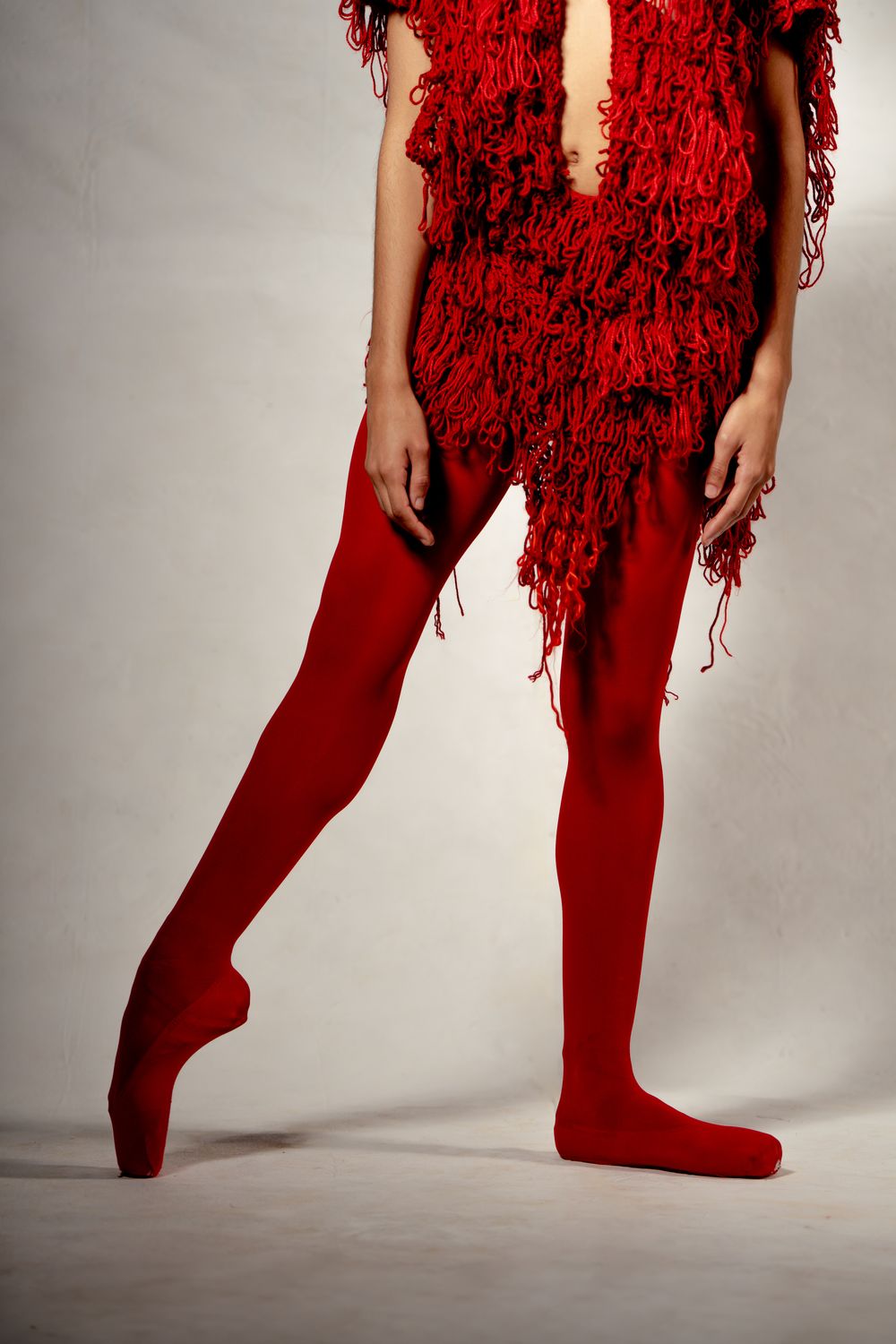 Model wearing red fringe dress with matching red tights and pointed toe shoes against gray background.
