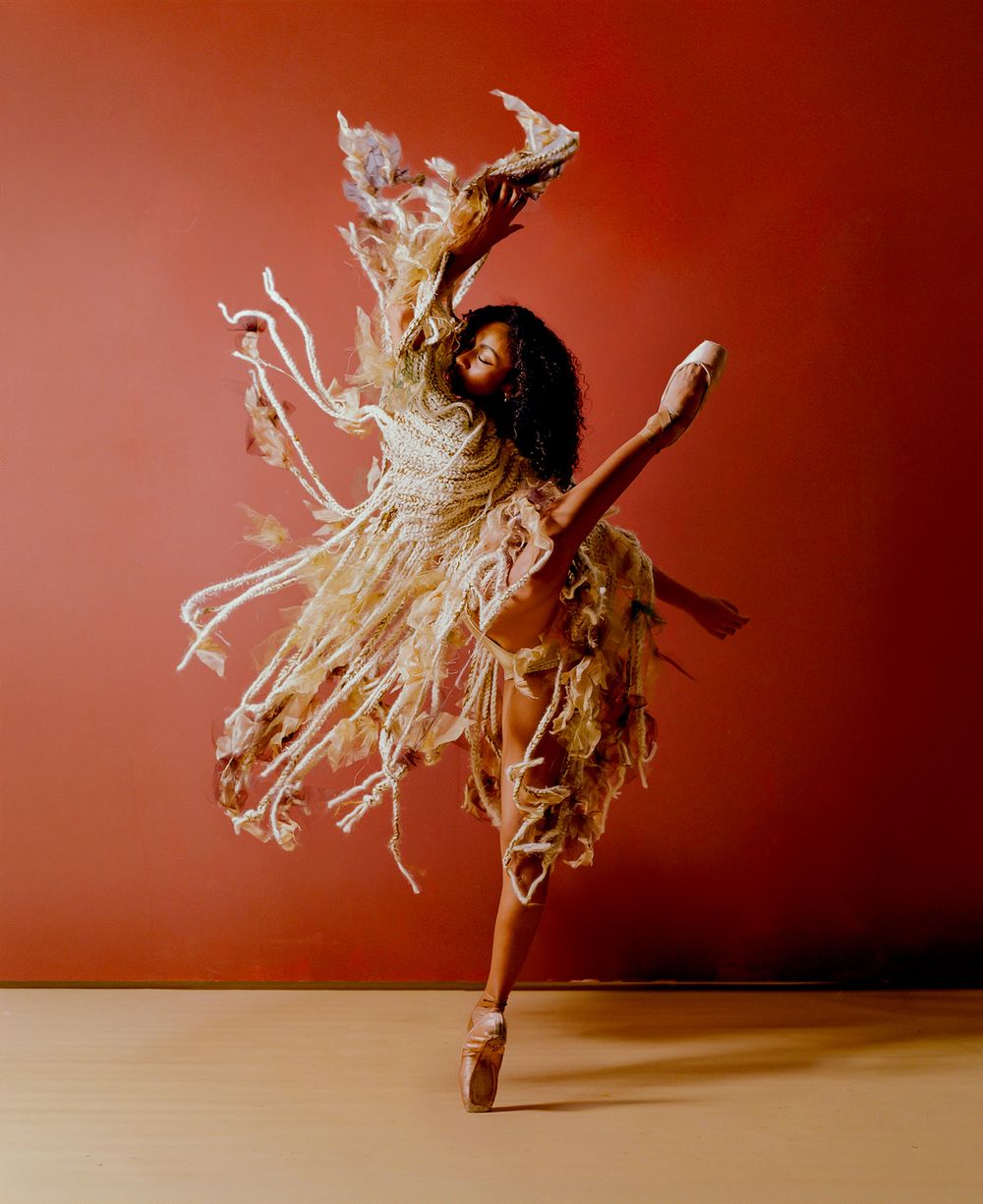 A ballet dancer in a flowing textured costume leaps gracefully against a warm red studio background.