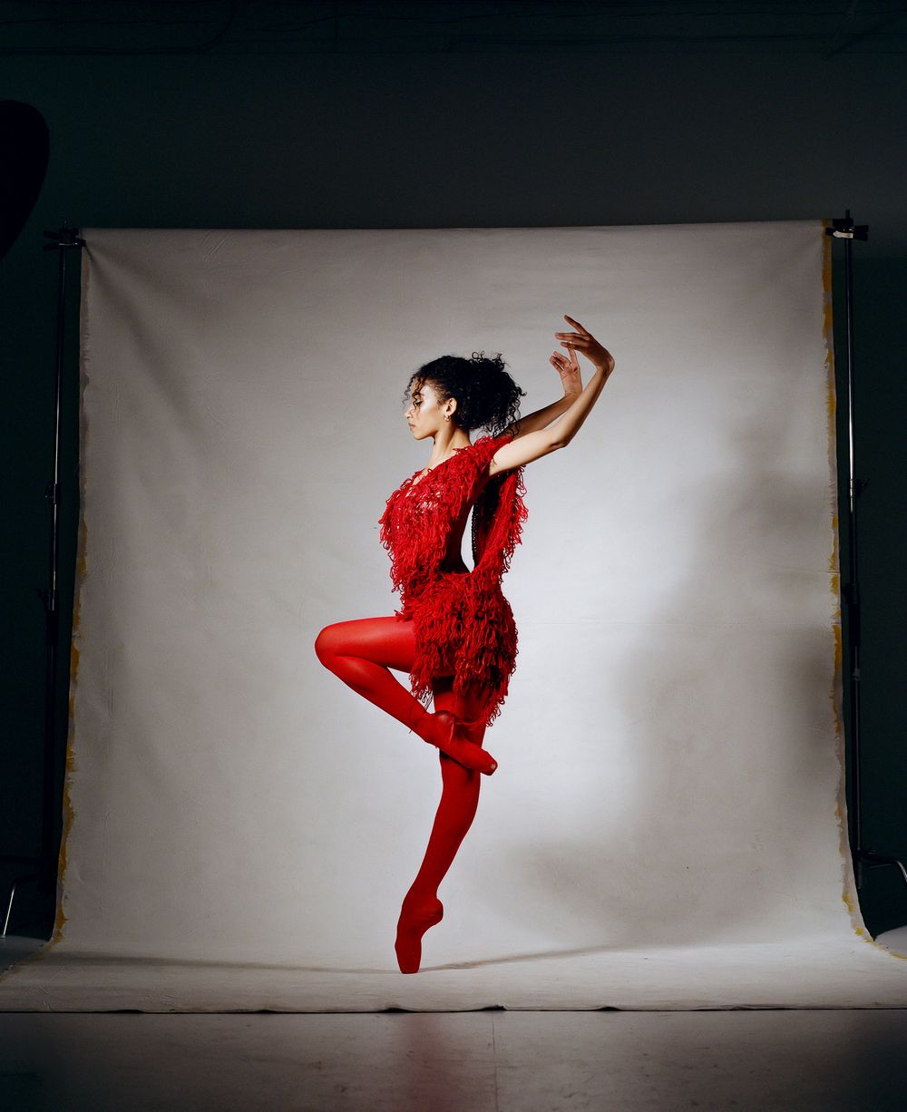 A dancer leaps in a vibrant red feathered costume and tights against a white backdrop.