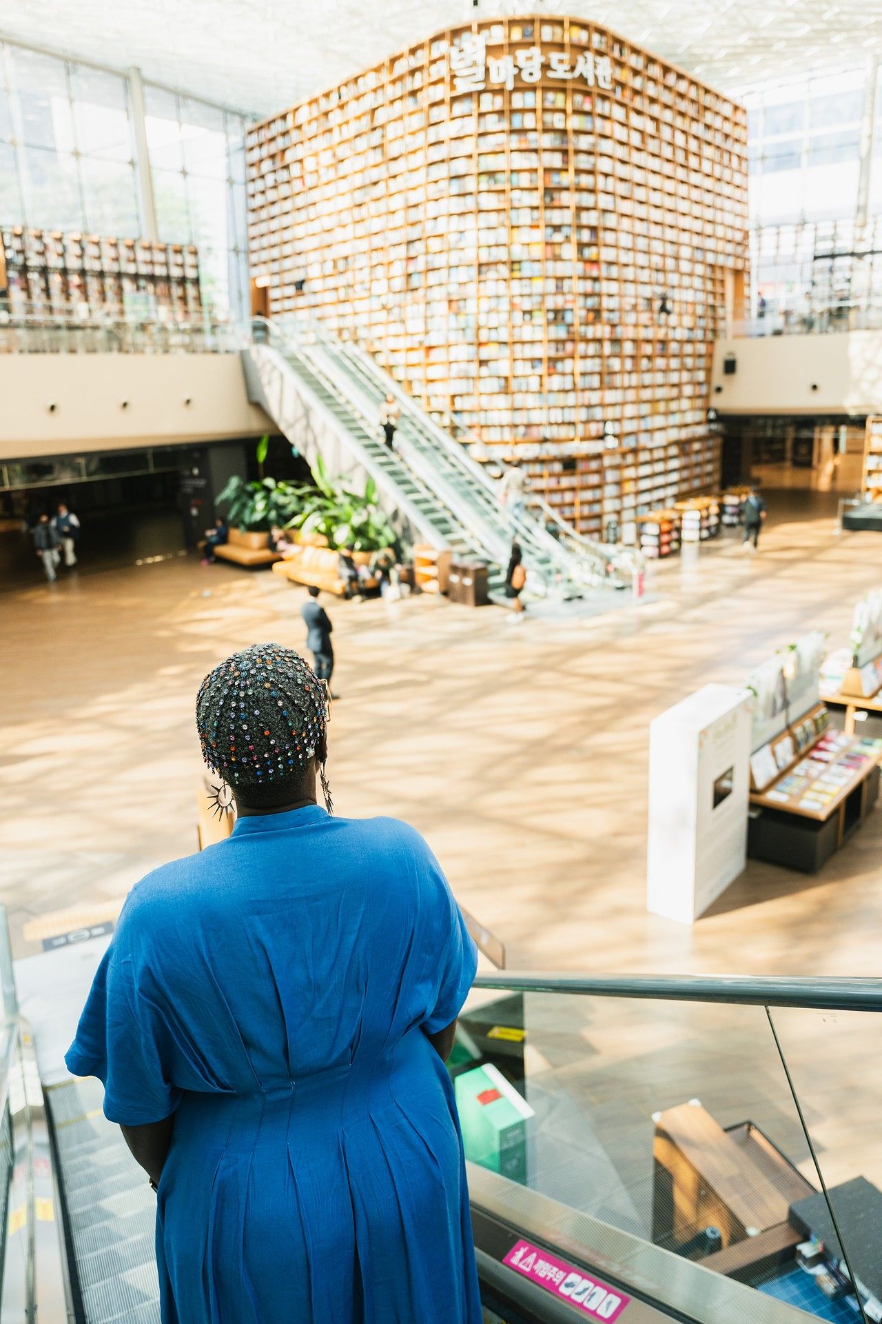 Woman in blue dress on escalator overlooking a large modern library with towering bookshelves.