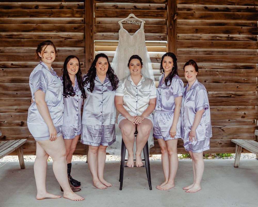Six bridesmaids in matching lavender satin pajamas pose together in front of a rustic wooden wall with a wedding dress hanging above.