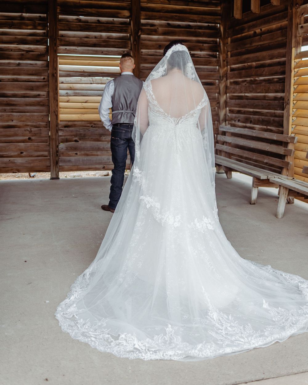 Bride in a white lace wedding gown with long veil and train stands with groom in rustic log barn.