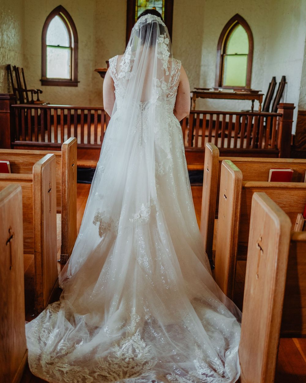 Bride in lace wedding gown with cathedral veil stands at church altar between wooden pews.