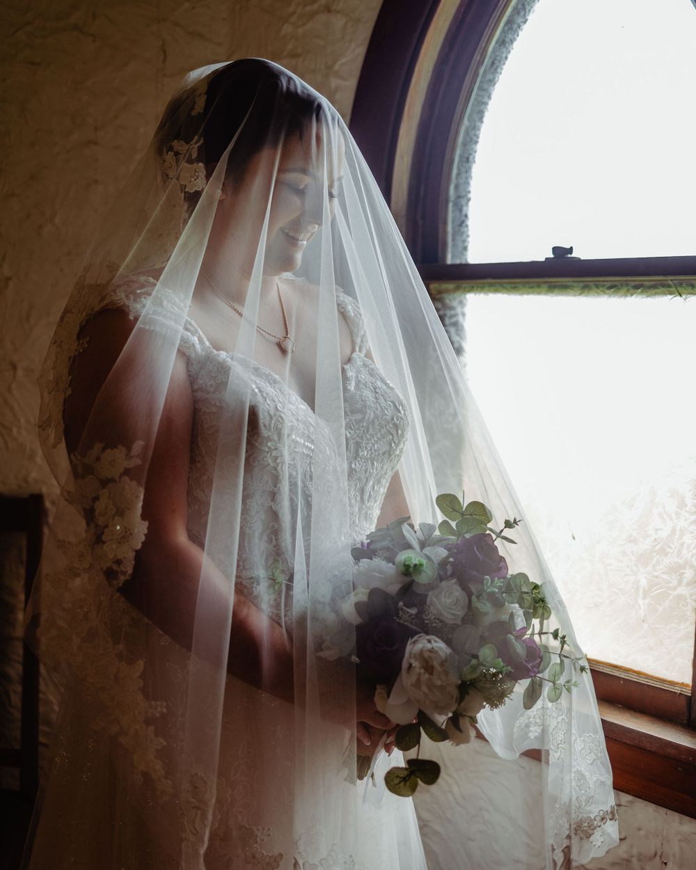 Bride in lace wedding dress and veil holds floral bouquet by arched window in soft natural light.