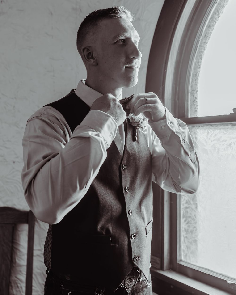 Black and white photo of a groom adjusting his boutonniere while wearing a vest and dress shirt near a window.