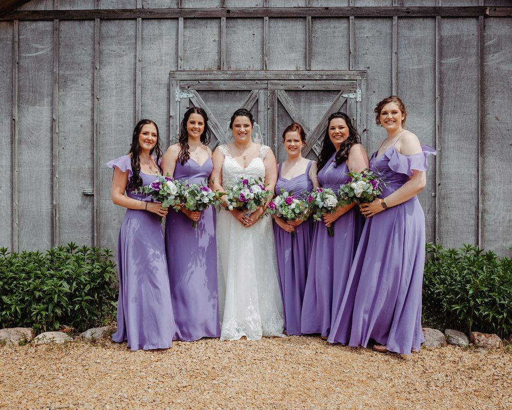 Bride in white gown poses with five bridesmaids in lavender dresses holding bouquets before a rustic barn.