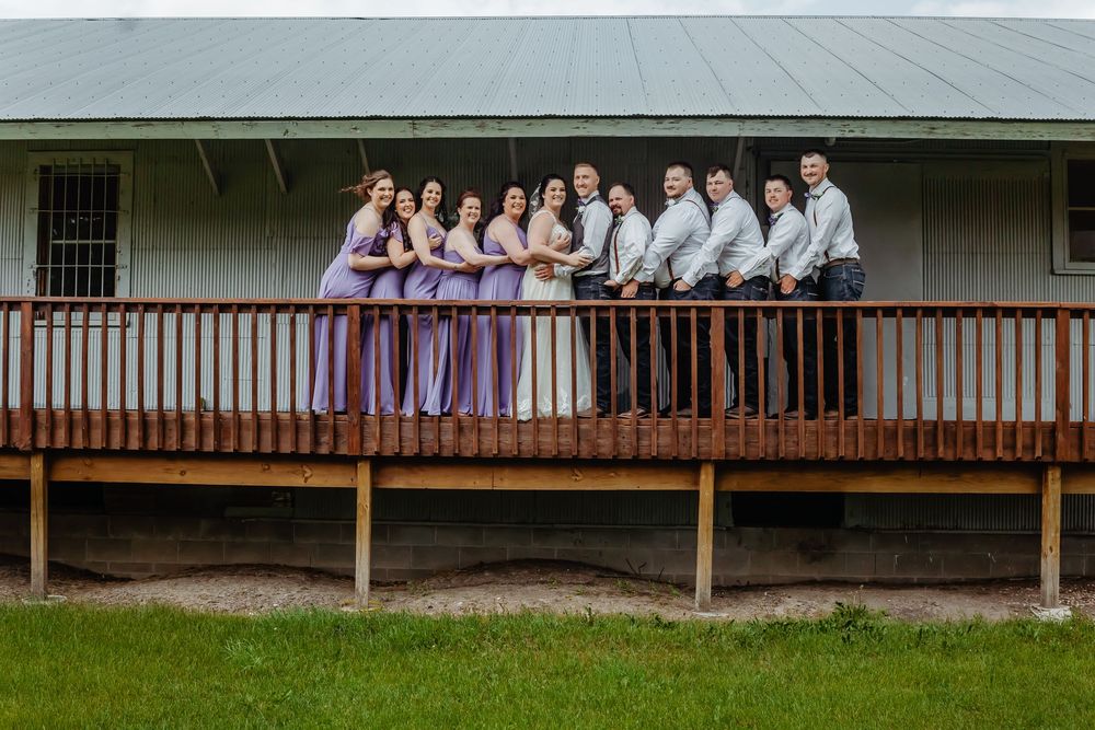 Wedding party poses on a wooden deck, bridesmaids in lavender gowns and groomsmen in gray suits beside the bride and groom.