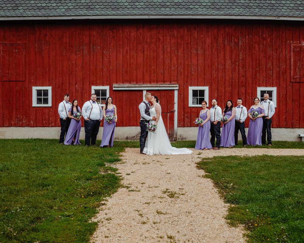 Wedding party poses before a red barn, bride and groom kissing at center flanked by groomsmen and bridesmaids.
