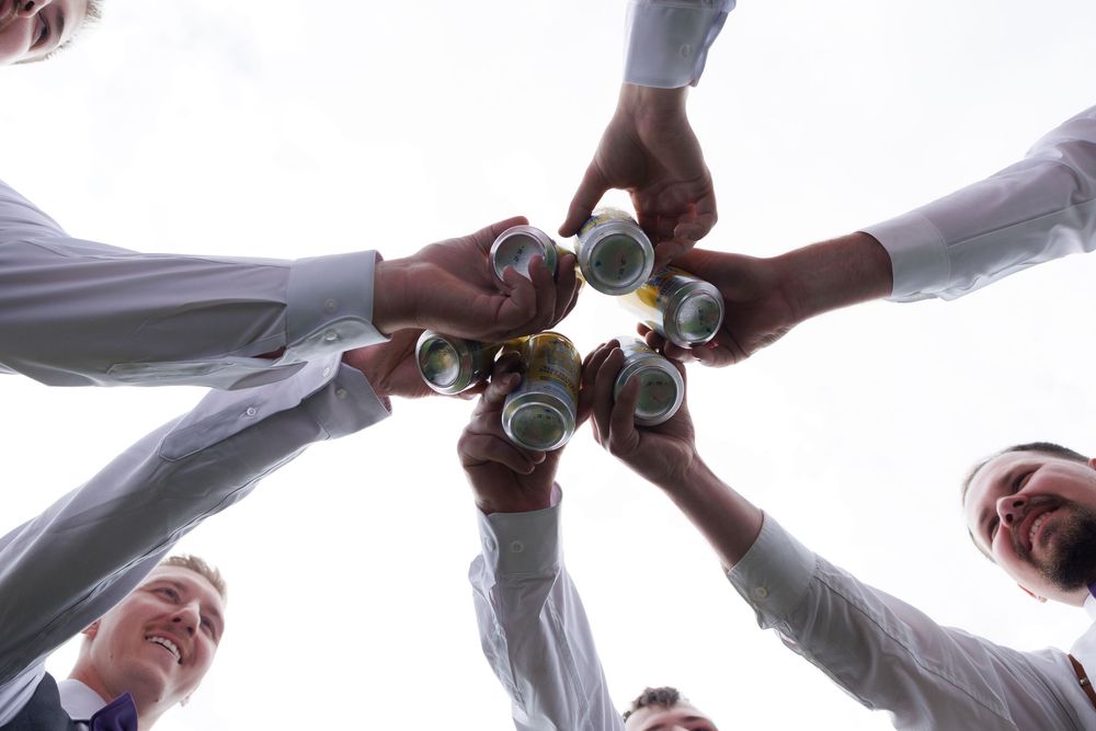 Group of groomsmen toasting with beer cans in a circle, viewed from below.