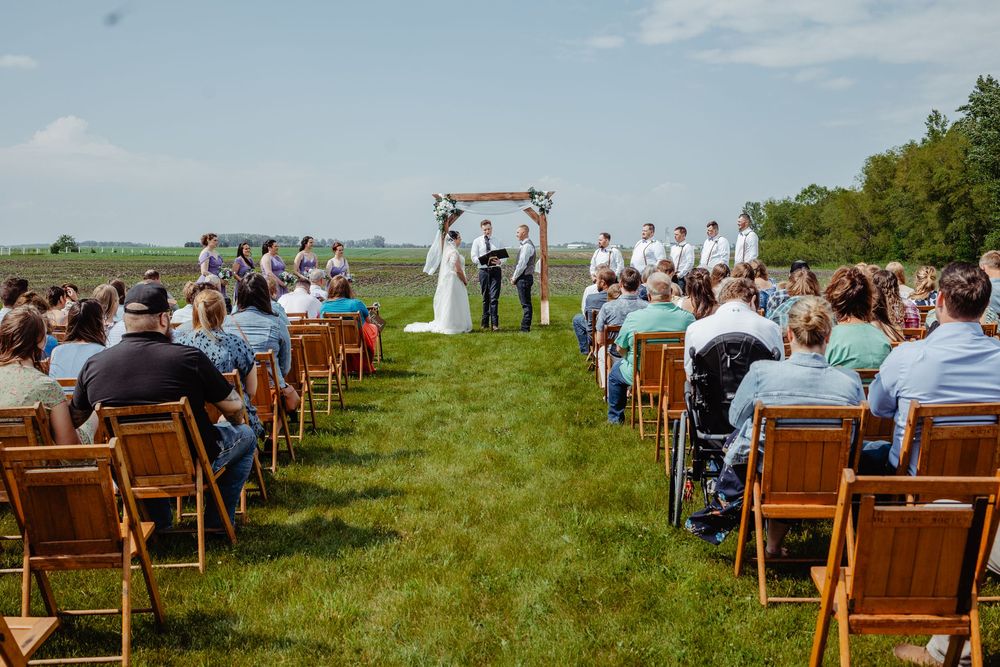 Outdoor wedding ceremony on a green lawn with guests seated in wooden chairs facing the altar under open sky.