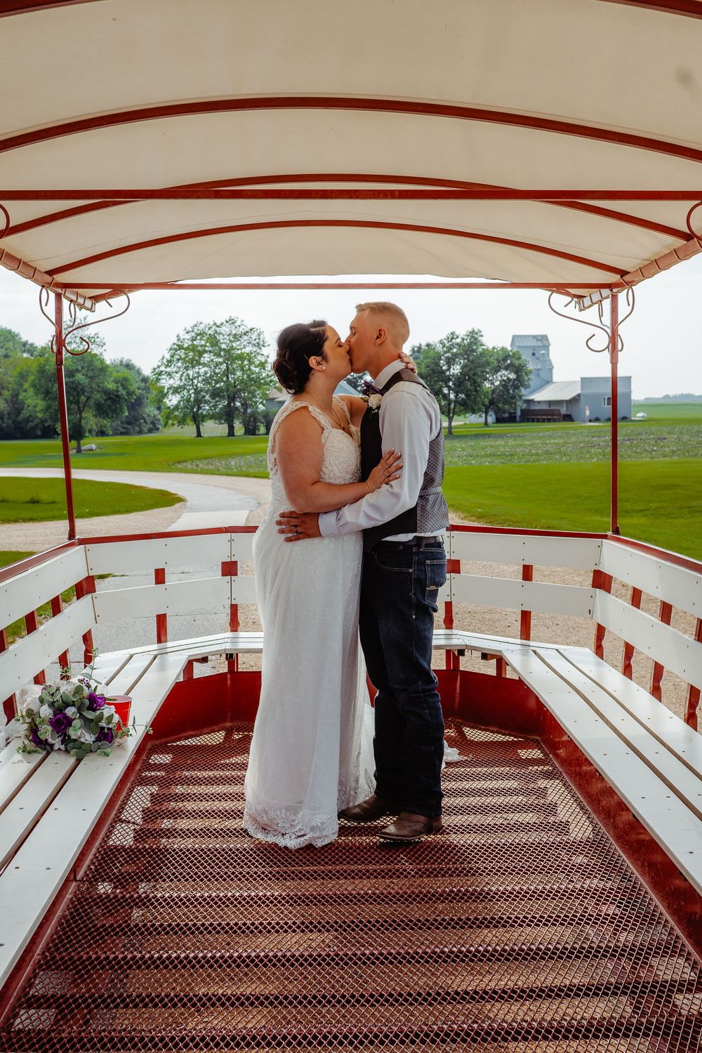 Bride and groom share a kiss inside a red and white covered wagon at an outdoor wedding venue.