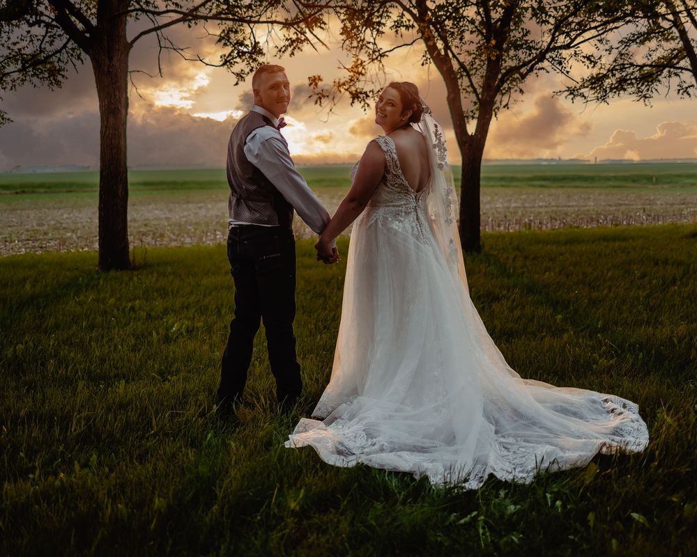 Bride and groom hold hands at sunset in green field with dramatic sky behind trees.