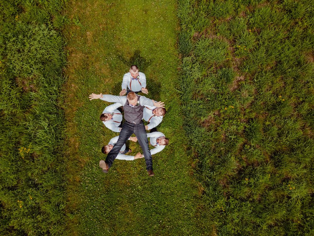 Aerial drone view of a groom and groomsmen lying in a star formation on green grass.