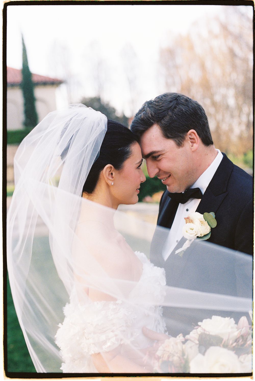 Bride and groom sharing an intimate moment, bride wearing veil and groom in black tuxedo with white boutonniere.