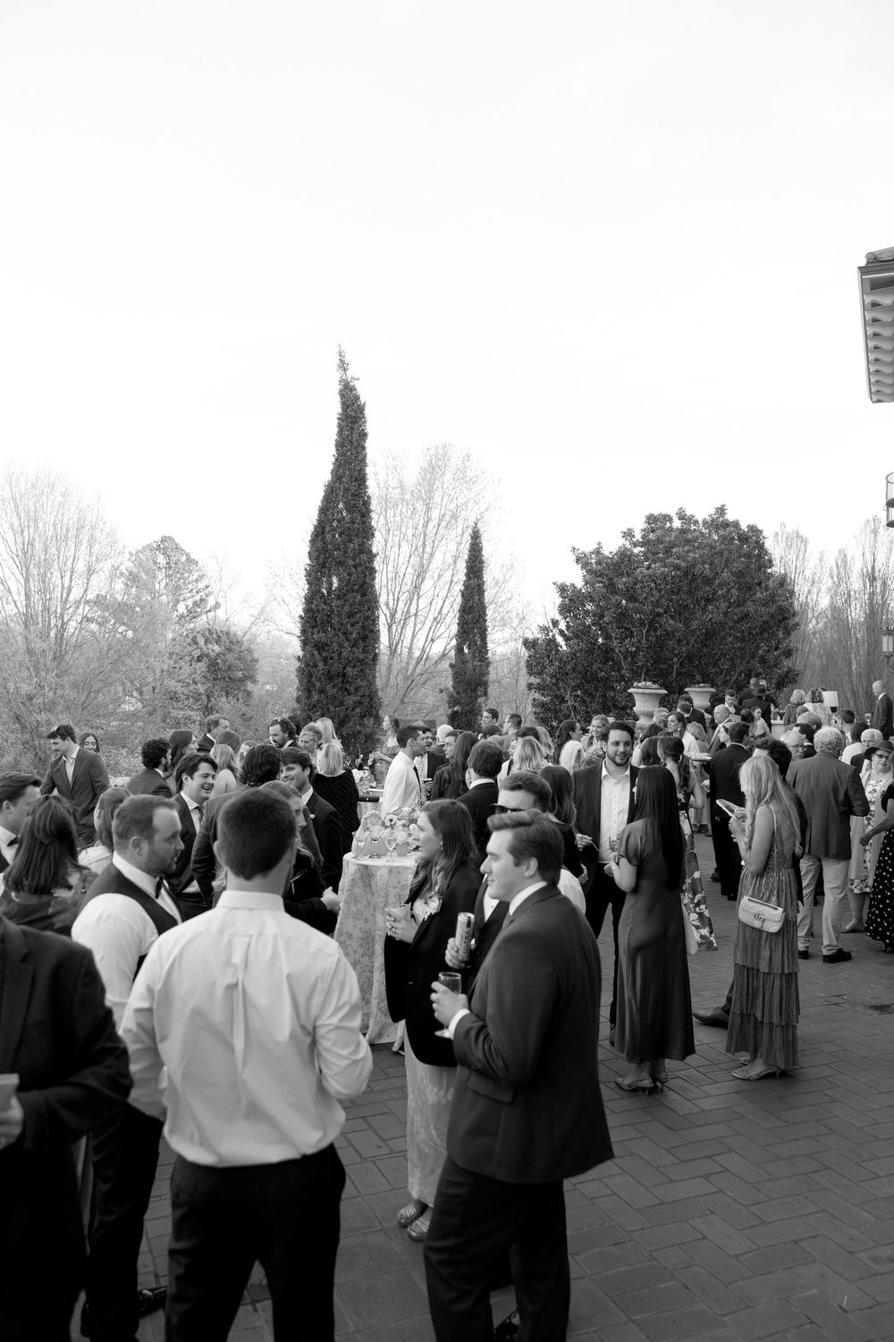 Black and white photo of elegantly dressed wedding guests mingling outdoors at a reception.