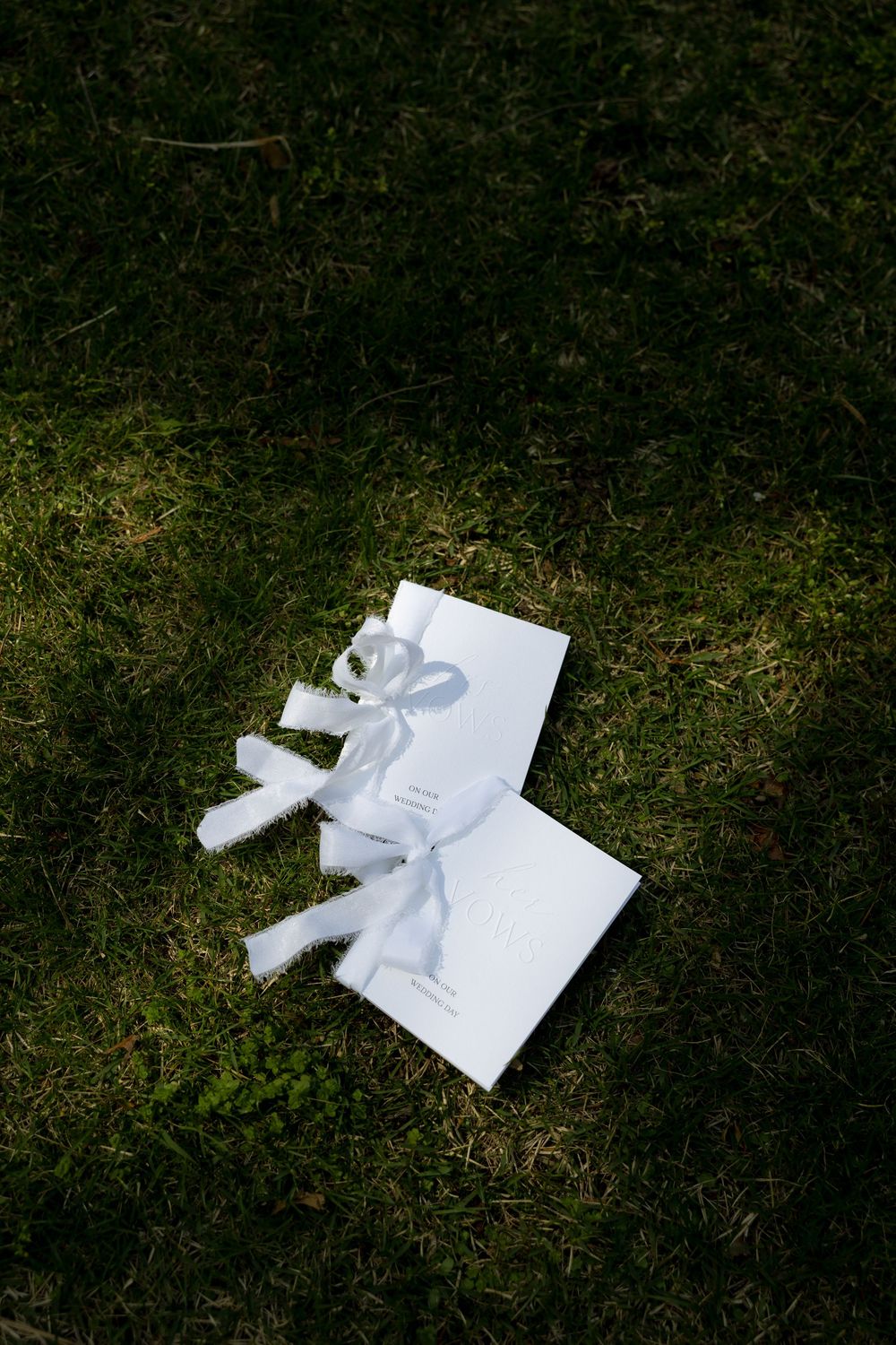Two white wedding vow books tied with ribbon bows resting on green grass.