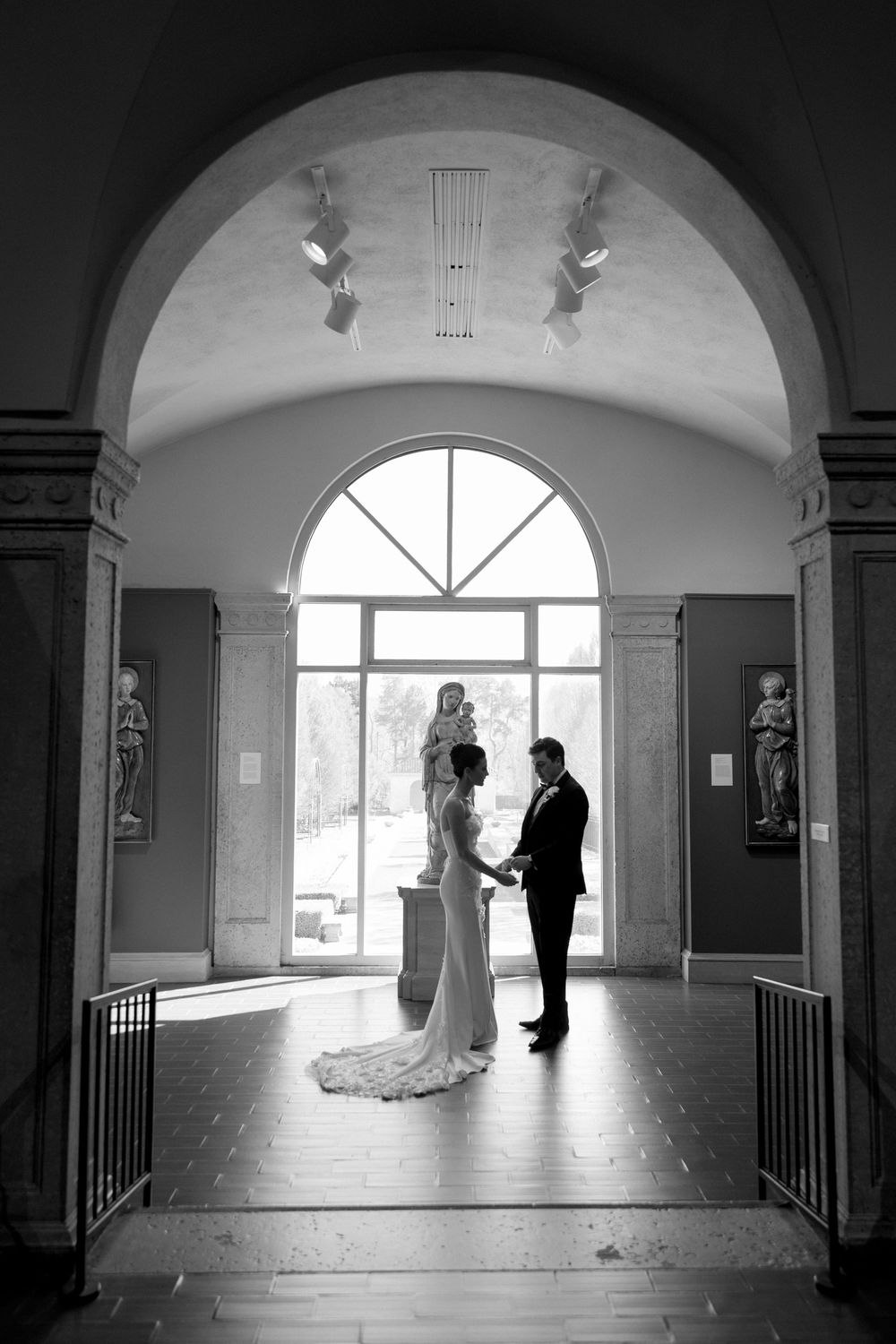 Bride and groom share an intimate moment under an arched doorway in an elegant museum hall, black and white photo.