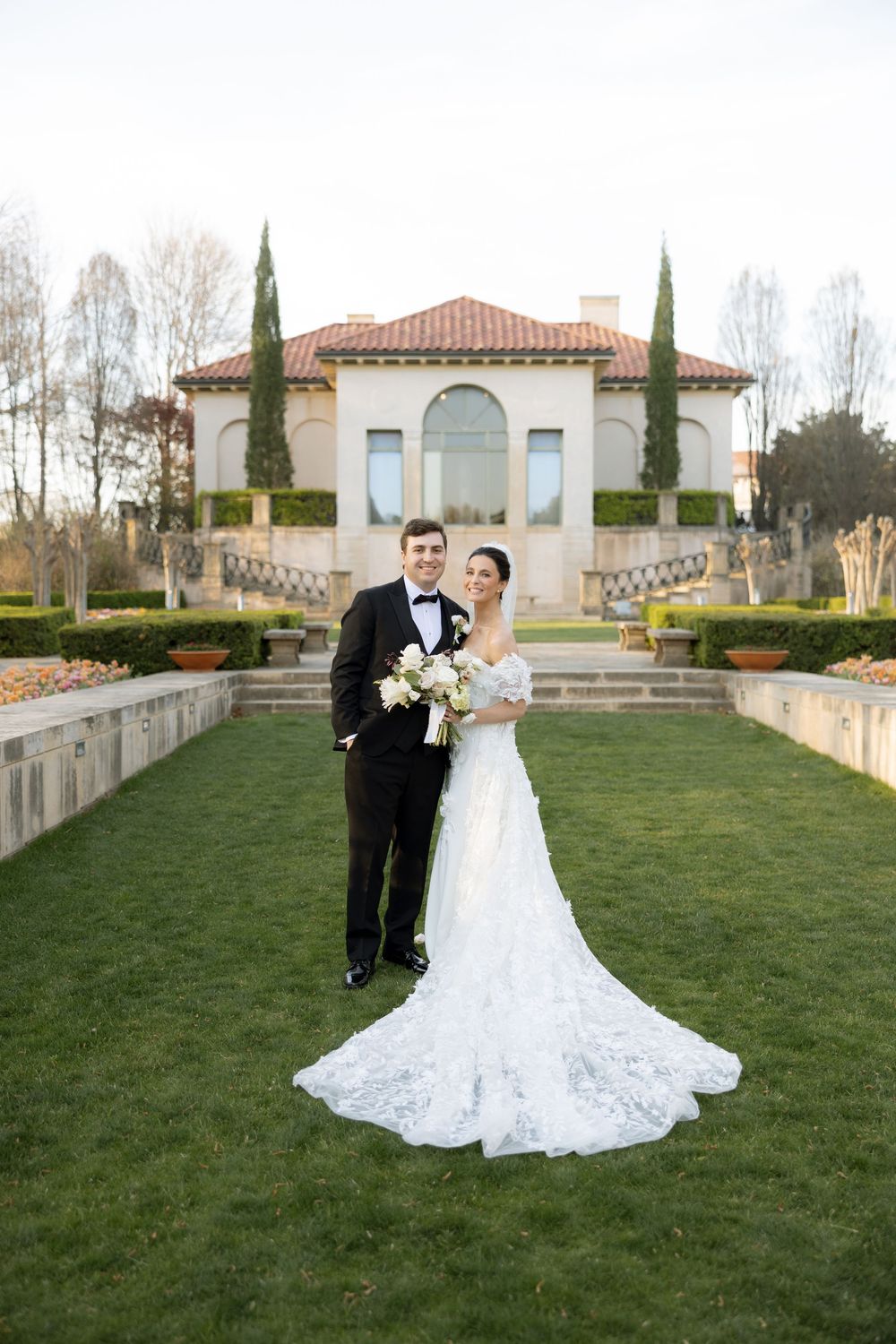 Bride and groom posing together on green lawn before elegant Italian-style villa with cypress trees.