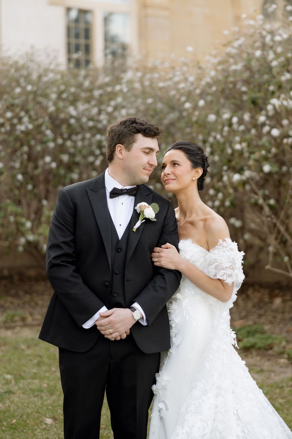 Bride in off-shoulder floral gown gazes at groom in black tuxedo standing before blooming white bushes.