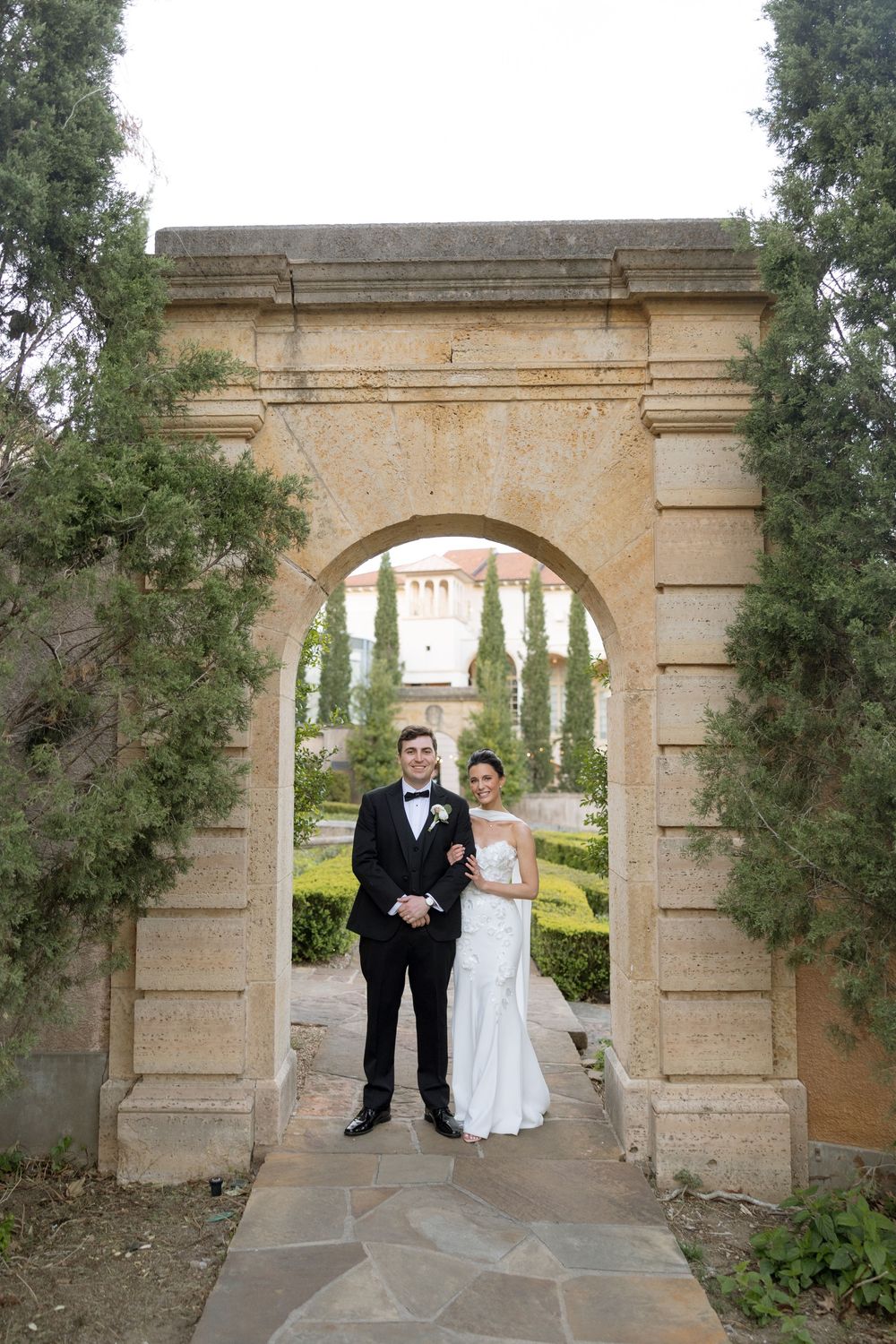Bride and groom posing under stone arch in formal garden at elegant wedding venue.