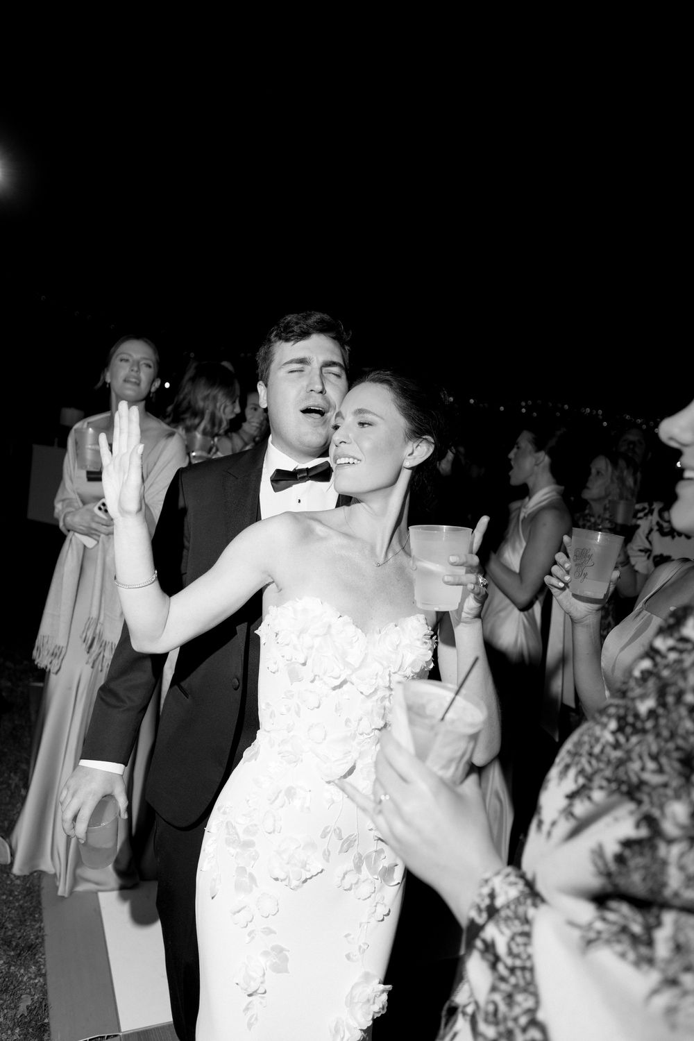 Bride and groom dance joyfully together at outdoor evening wedding reception in black and white.