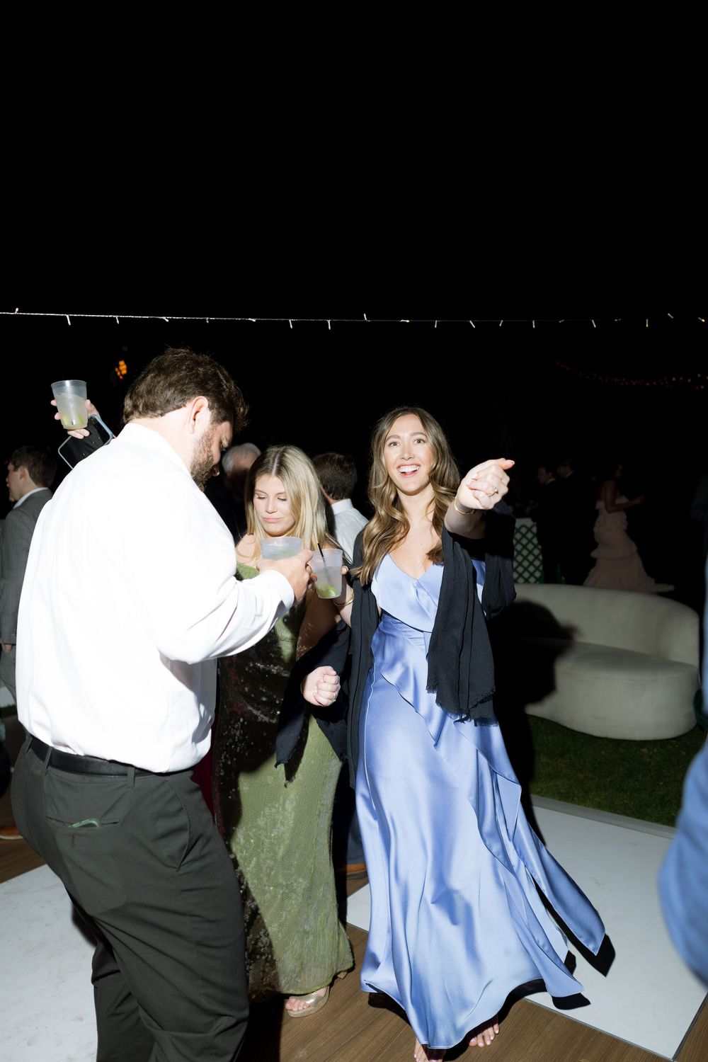 Wedding guest in blue satin gown dancing and pointing at outdoor evening reception under string lights.