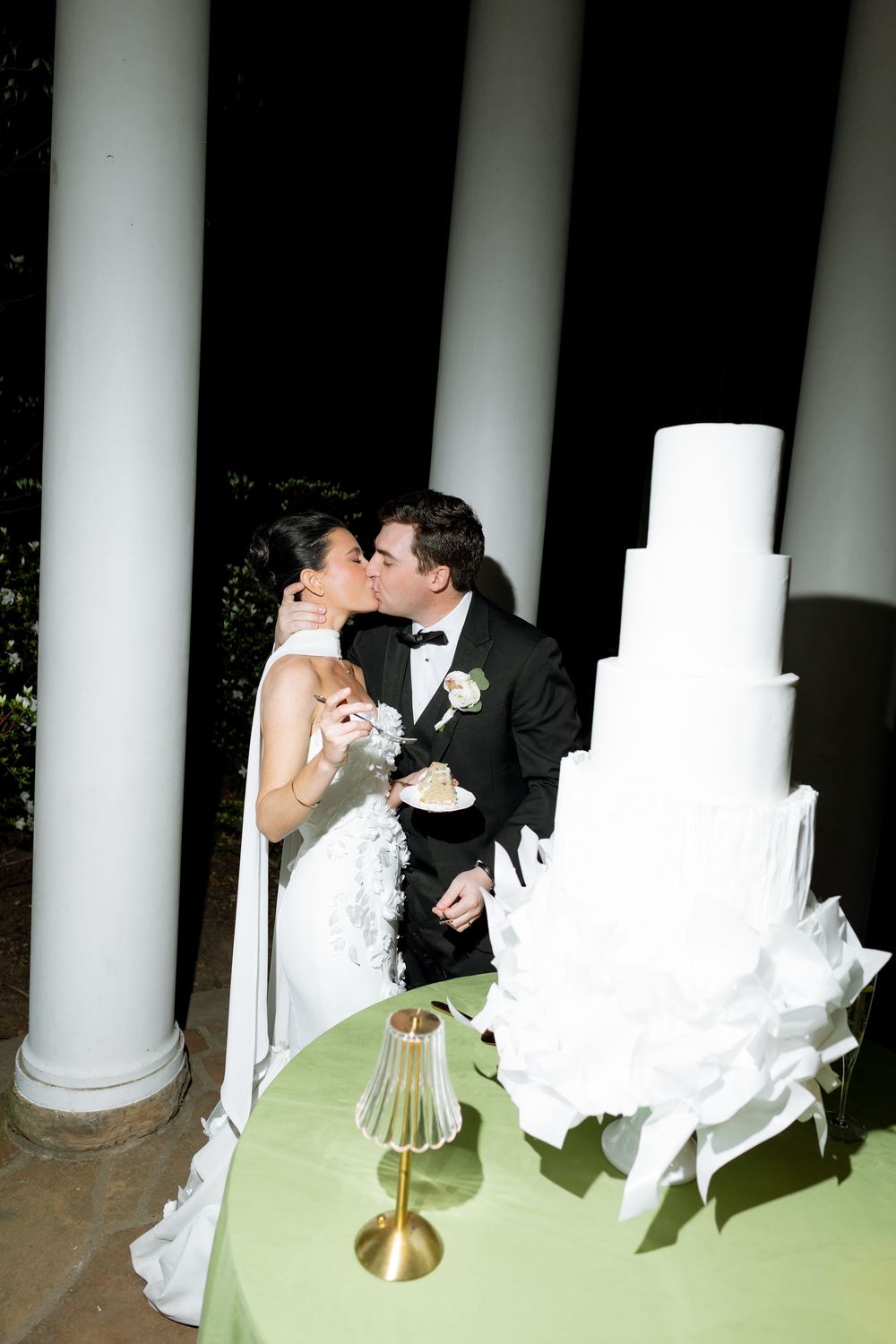 Bride and groom sharing a kiss beside tall white ruffled wedding cake between white columns at night.