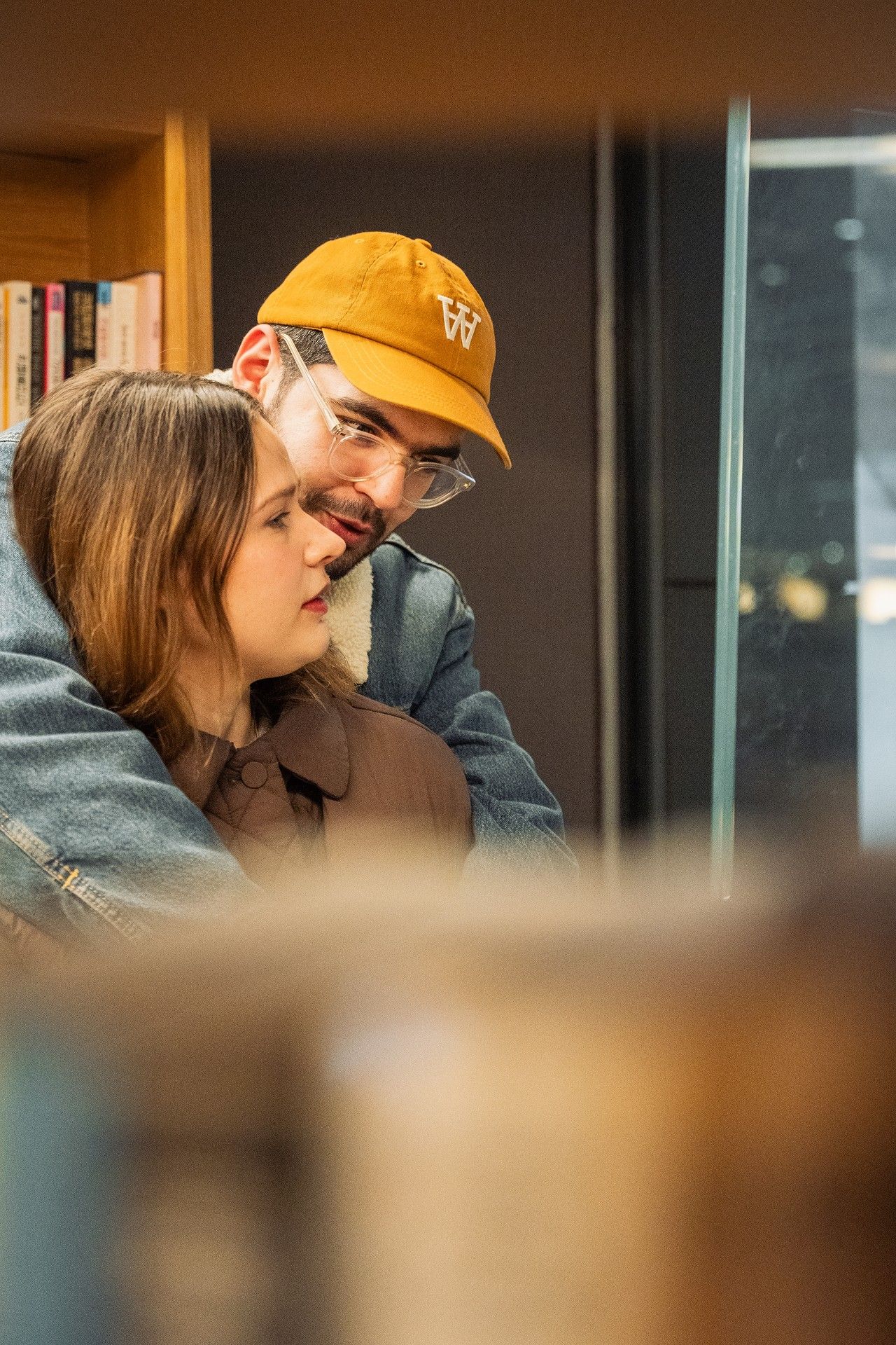 A couple shares an intimate moment, man in yellow cap embracing woman near a bookshelf indoors.