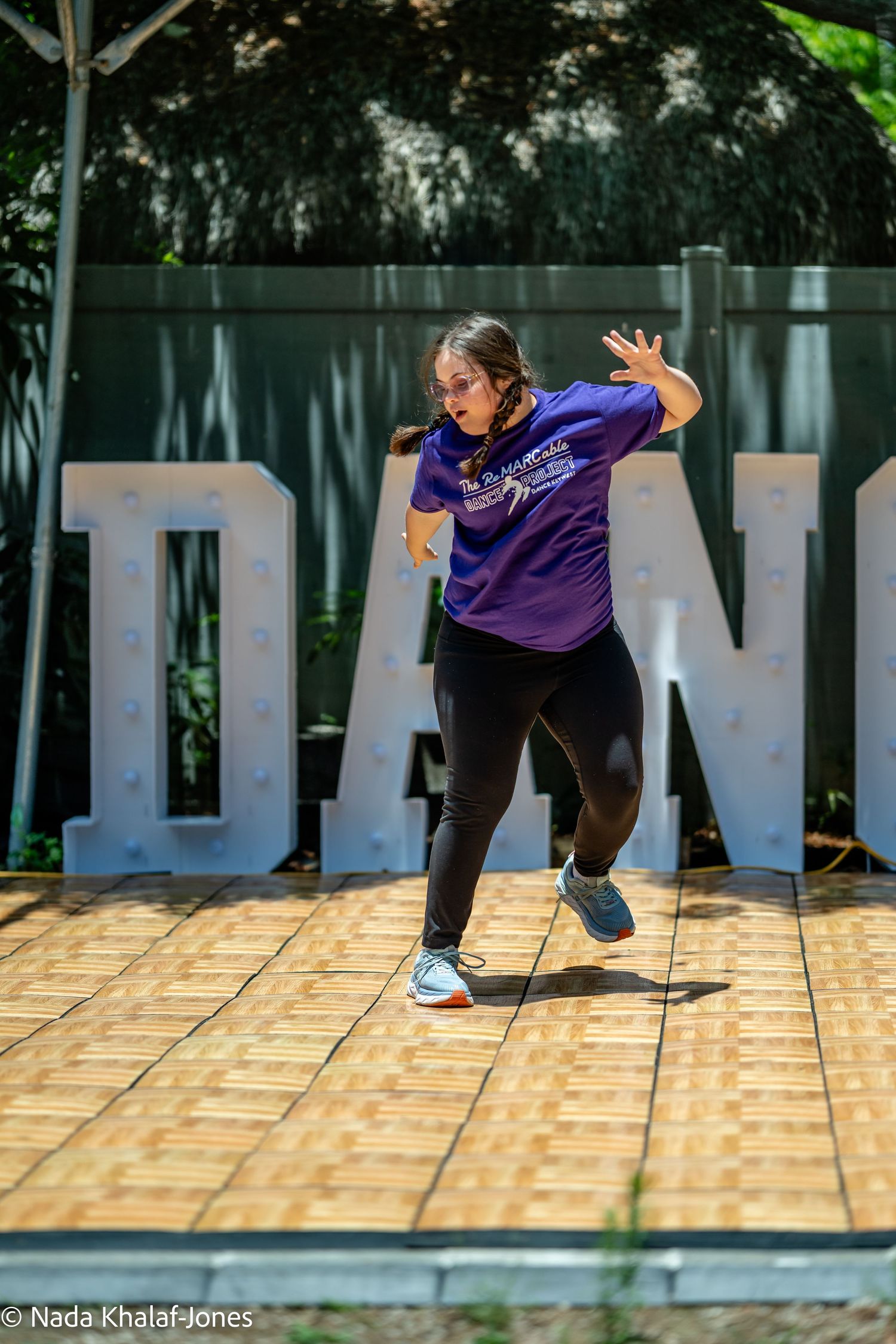 A young woman in a purple t-shirt performs an expressive dance move outdoors in front of large DANCE letters.