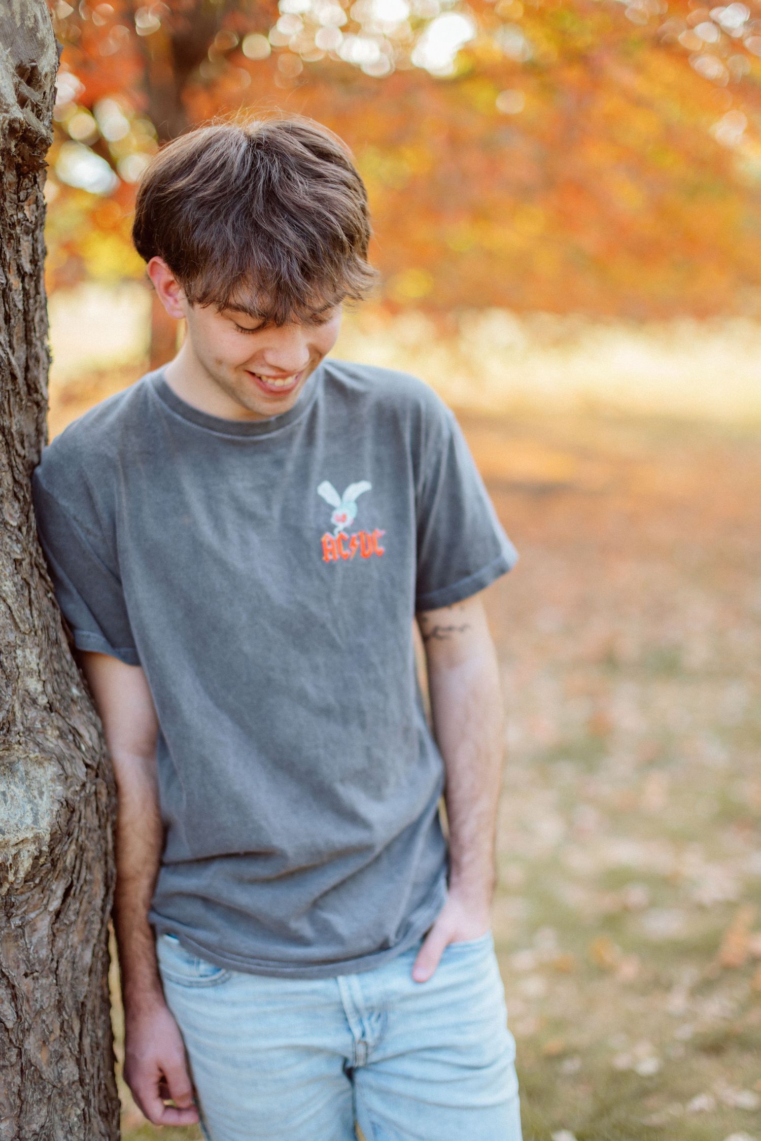 A high school senior boy leans casually against a tree with hands in his pockets, smiling and looking downward at the autumn leaves.