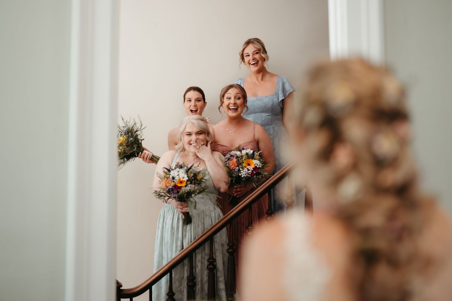 Bridesmaids in blue and orange dresses holding bouquets react joyfully on a staircase as bride approaches.