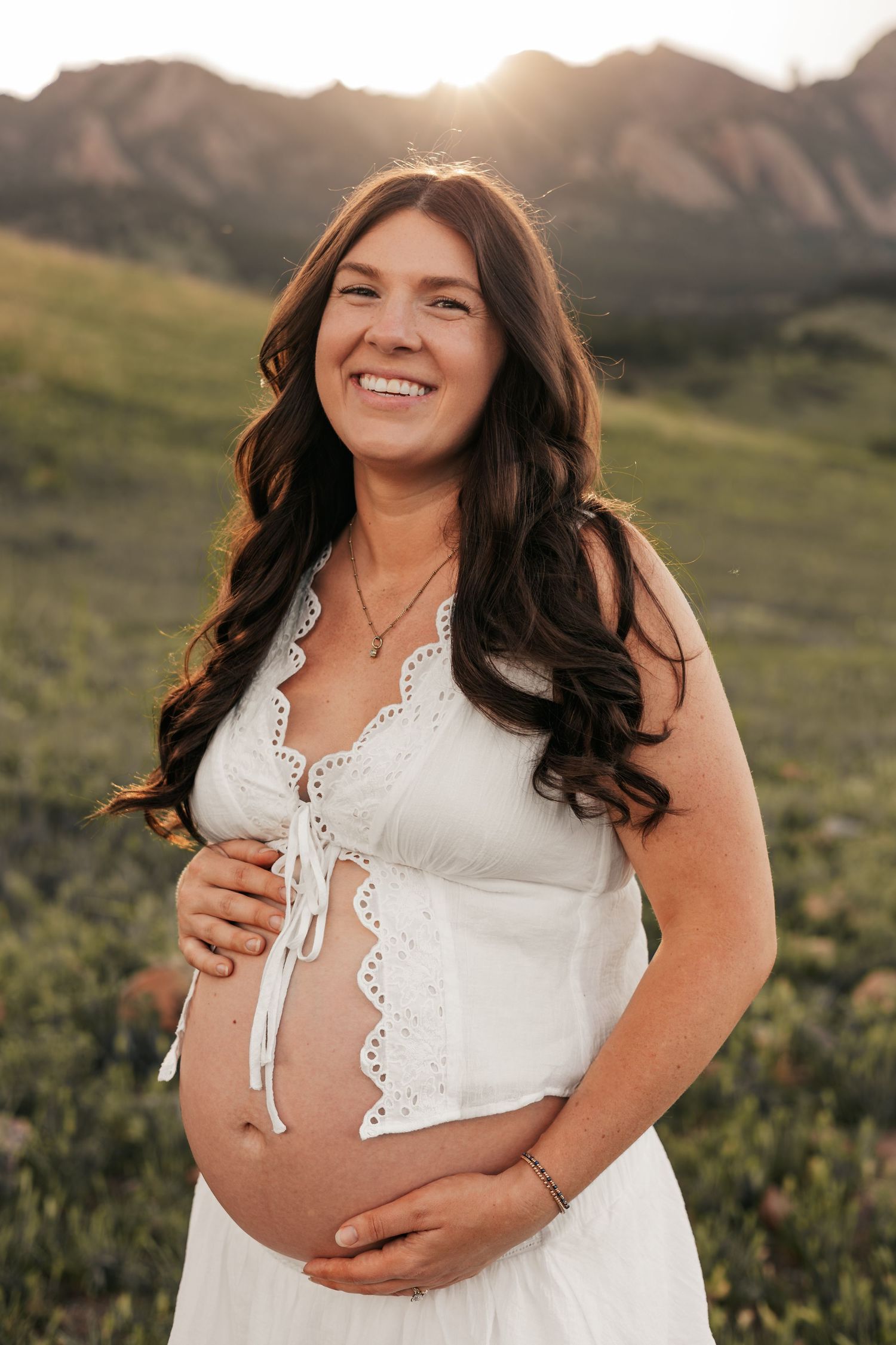 Smiling pregnant woman in white dress cradling baby bump in scenic mountain meadow at golden hour sunset | Boulder family photographer