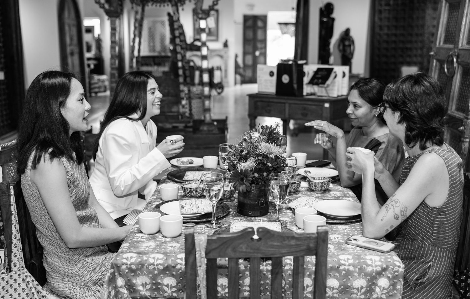 Four women enjoy a lively conversation at a decorated dining table in a cozy, eclectic interior setting.