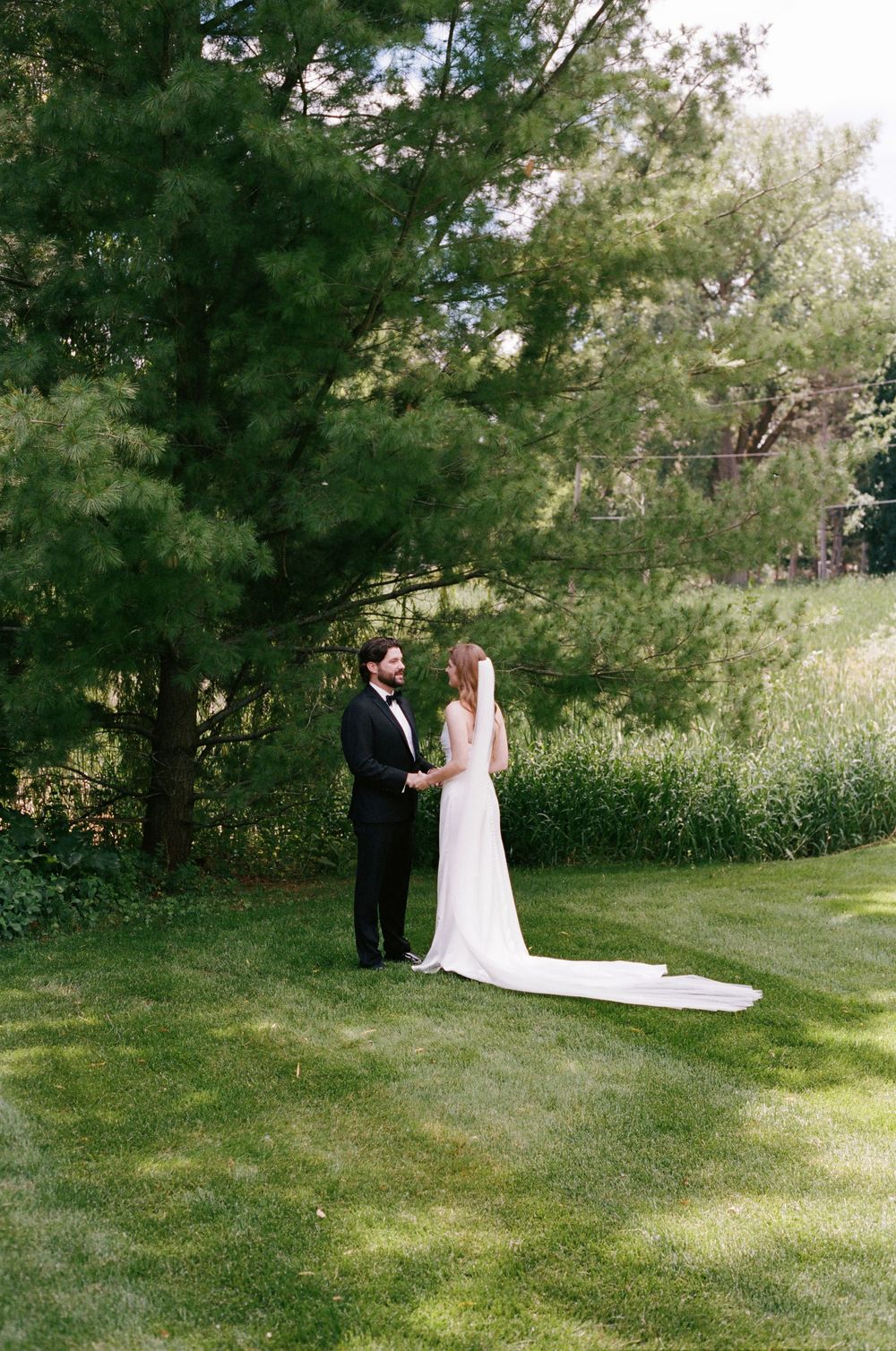 Bride and groom share a romantic moment on a lush green lawn surrounded by tall trees.