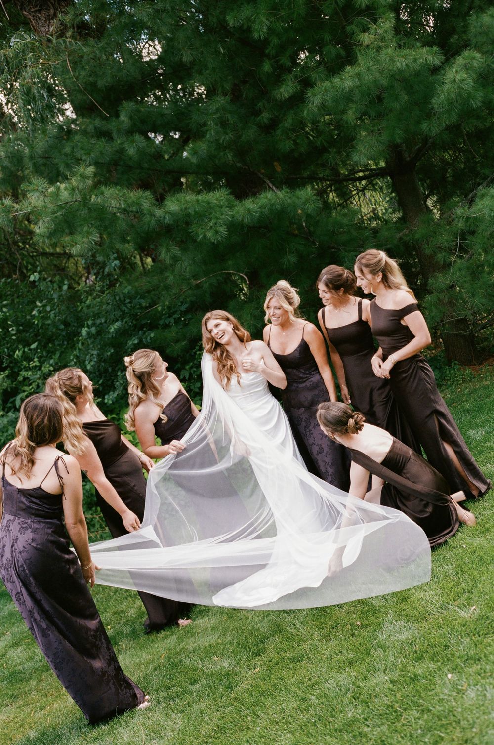 Bride in white gown with bridesmaids in black dresses arranging flowing veil on green lawn outdoors.