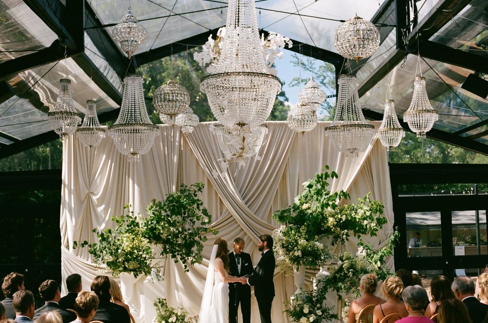 Elegant wedding ceremony under crystal chandeliers with draped white fabric and lush greenery altar.