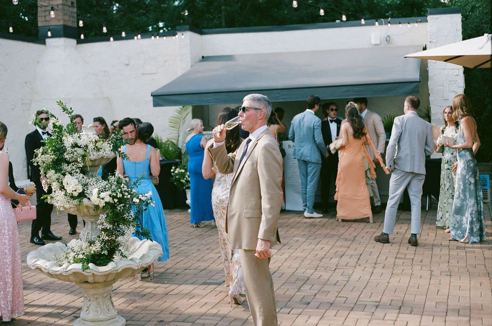 Man in tan suit enjoys outdoor wedding cocktail hour on brick patio with floral arrangements.