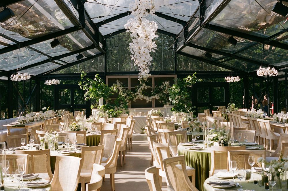 Stunning glass-ceiling wedding reception hall with chandelier, round tables, and greenery centerpieces.