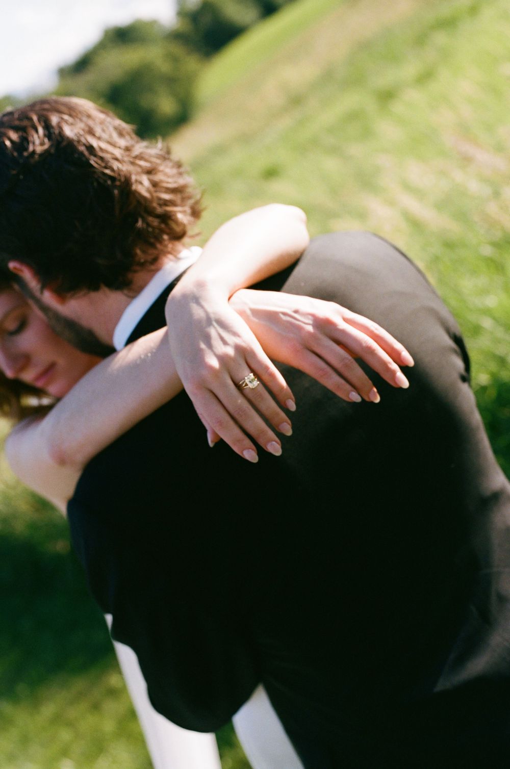 Bride embracing groom from behind outdoors, showcasing diamond wedding ring against his black suit jacket.