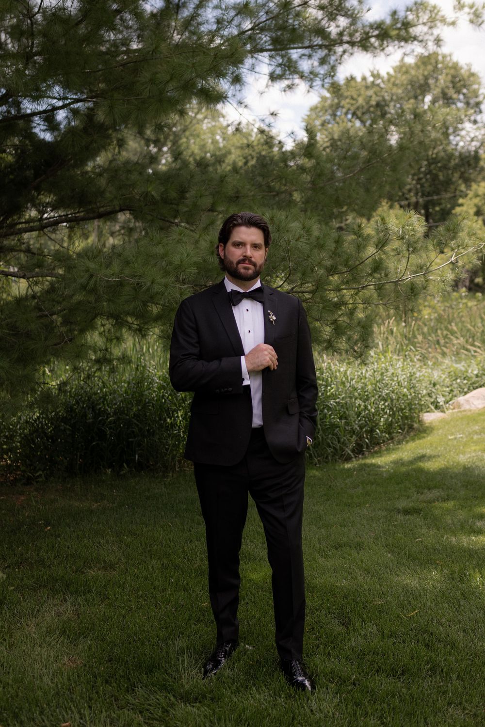 Dapper groom in black tuxedo with bow tie posing confidently outdoors among lush green trees and lawn.