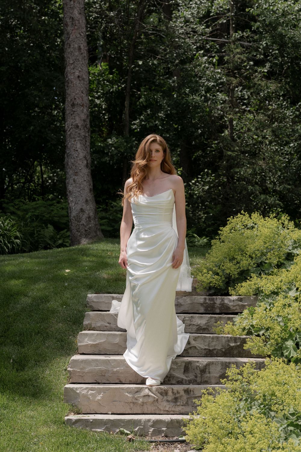Elegant bride in a white strapless satin gown descending stone garden steps surrounded by lush greenery.