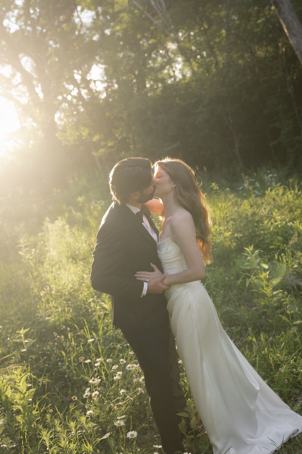 Bride and groom sharing a romantic kiss in a sunlit forest clearing, bride in white gown, groom in black tuxedo.