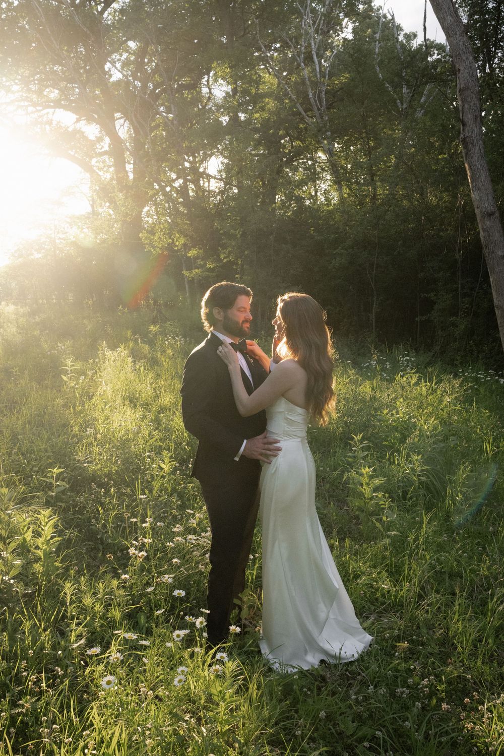 Wedding couple holding each other tenderly in a golden sunlit meadow, groom in tuxedo and bride in white dress.