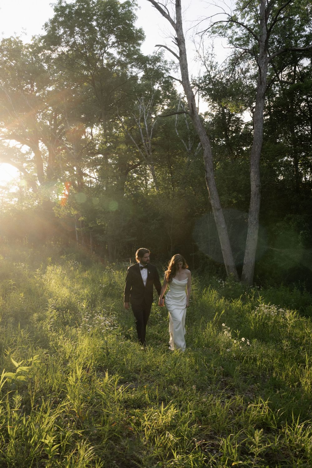 Newlywed couple holding hands strolling through green meadow with golden sunset light filtering through trees.