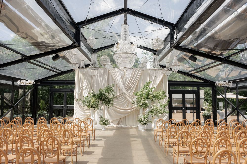 Elegant greenhouse wedding ceremony with crystal chandelier, draped ivory fabric backdrop, lush greenery, and wooden cross-back chairs.