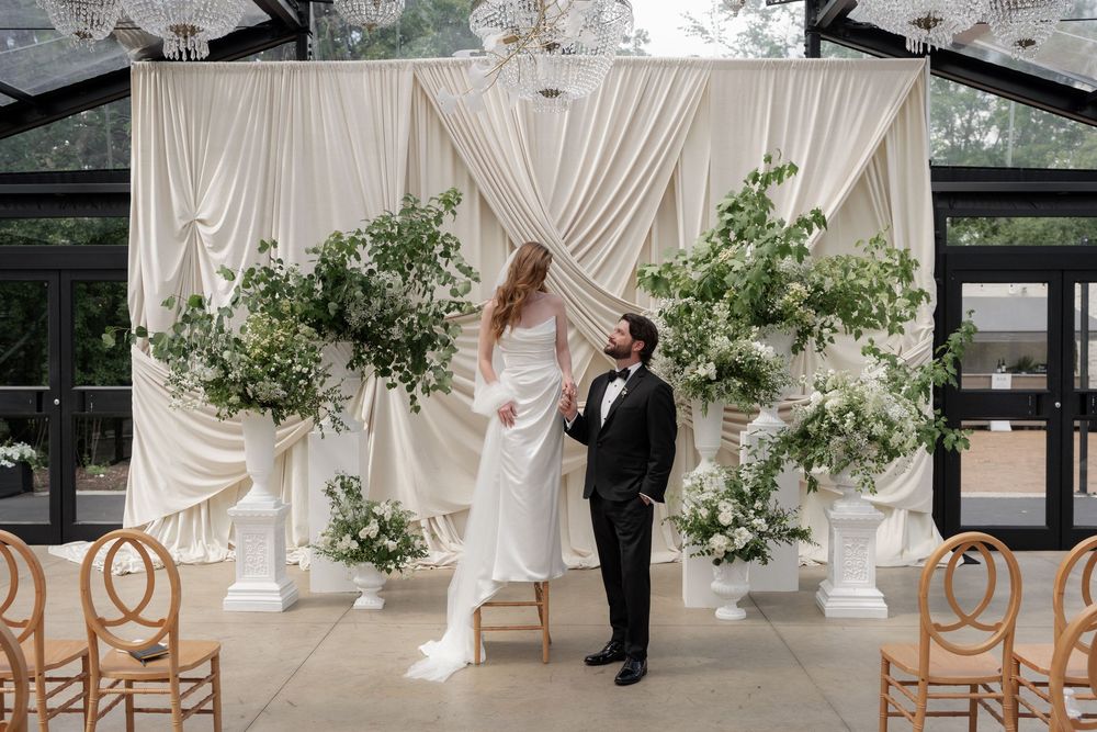 Bride and groom stand beneath a stunning white draped altar adorned with lush greenery in a glass venue.
